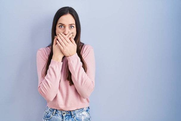 Young brunette woman standing over blue background shocked covering mouth with hands for mistake. secret concept.  - Photo, image