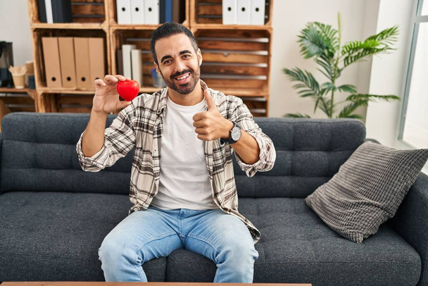 Jonge Spaanse man met baard die werkt aan liefdestherapie op consultatiebureau glimlachend vrolijk en positief, duim omhoog doen uitstekend en goedkeuring teken  - Foto, afbeelding