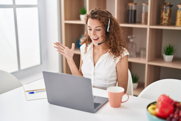 Young caucasian woman wearing call center agent headset working from home celebrating achievement with happy smile and winner expression with raised hand  - Photo, Image