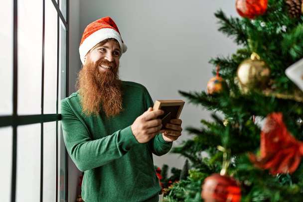 Young redhead man looking photo standing by christmas tree at home - Foto, Bild