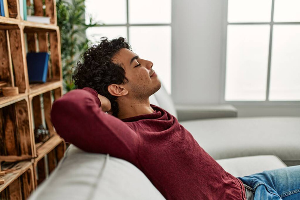 Young hispanic man relaxing with hands on head sitting on the sofa at home. - Foto, imagen