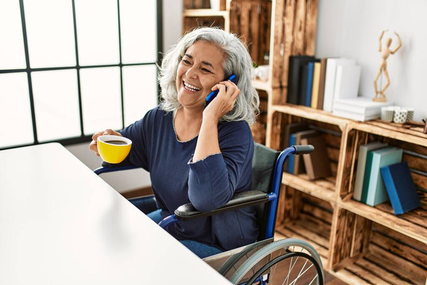 Középkorú, ősz hajú, fogyatékos nő, aki az okostelefonon beszél, és otthon tolószéken ülve kávézik.. - Fotó, kép