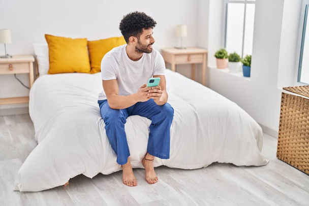 Nuori arabi mies käyttää älypuhelinta istuu sängyssä makuuhuoneessa - Valokuva, kuva