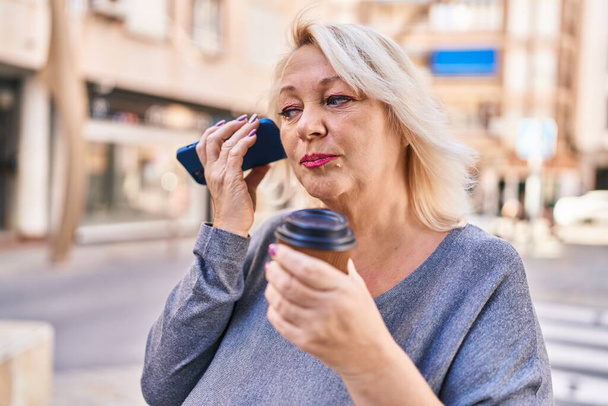 Middle age blonde woman listening audio message by the smartphone drinking coffee at street - Zdjęcie, obraz