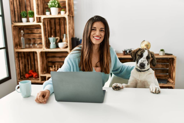 Jonge vrouw met laptop zittend op tafel met hond thuis - Foto, afbeelding