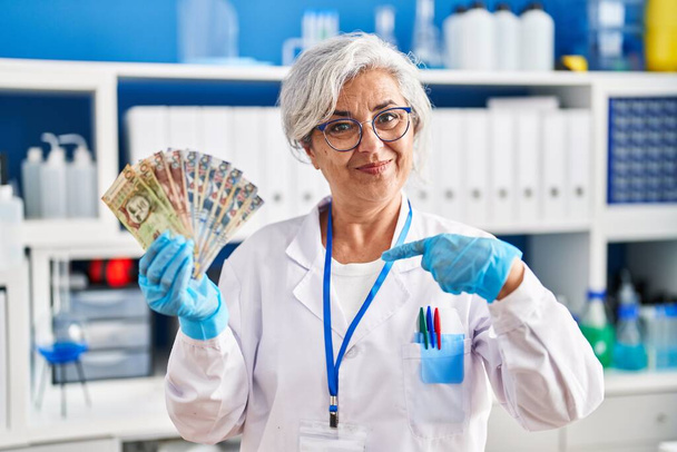 Middle age woman with grey hair working at scientist laboratory holding polish zloty banknotes pointing finger to one self smiling happy and proud  - 写真・画像