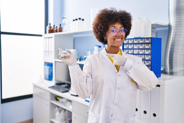 Junge Afroamerikanerin, die im wissenschaftlichen Labor arbeitet, lächelt und blickt in die Kamera, die mit zwei Händen und Fingern zur Seite zeigt.  - Foto, Bild