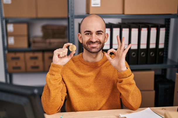 Молодой лысый человек с бородой работает в электронной коммерции малого бизнеса держа биткойн делает хорошо знак с пальцами, улыбаясь дружелюбный жест отличный символ  - Фото, изображение