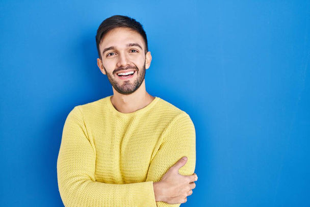 Een Spaanse man die over een blauwe achtergrond staat te glimlachen met gekruiste armen en naar de camera kijkt. positieve persoon.  - Foto, afbeelding