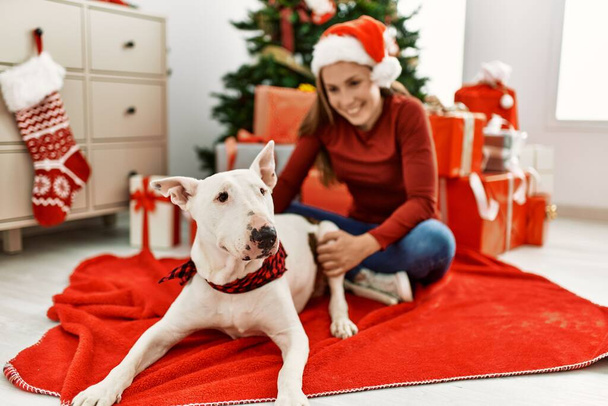 Молода біла жінка посміхається впевнено сидячи з собакою на ялинку вдома
 - Фото, зображення