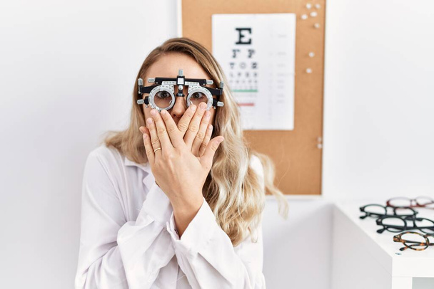 Young beautiful optician woman wearing optometry glasses at the clinic shocked covering mouth with hands for mistake. secret concept.  - Foto, imagen