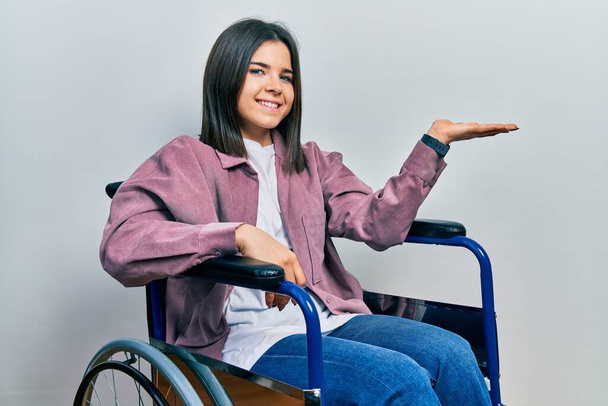 Young brunette woman sitting on wheelchair smiling cheerful presenting and pointing with palm of hand looking at the camera.  - Фото, изображение