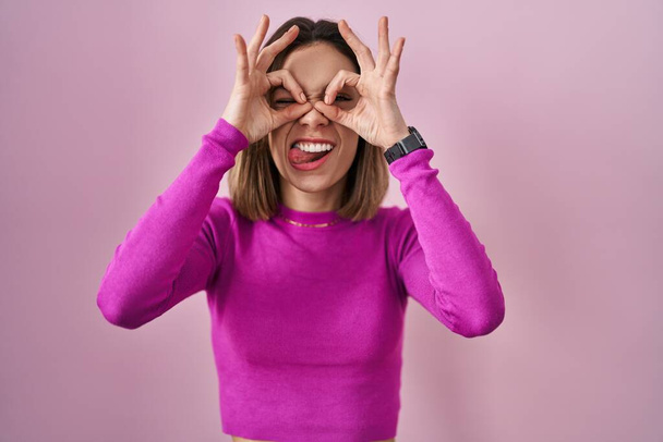 Mujer hispana de pie sobre fondo rosa haciendo un buen gesto como prismáticos sacando la lengua, ojos mirando a través de los dedos. expresión loca.  - Foto, Imagen
