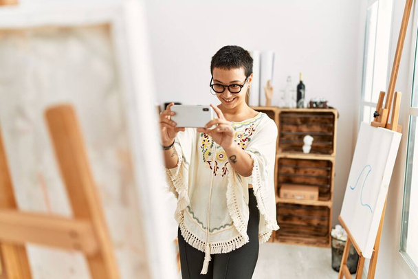 Молода іспаномовна жінка робить фотографію за допомогою смартфона на полотні малює в художній студії
 - Фото, зображення