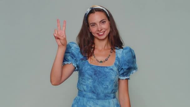 Lovely happy young woman showing victory sign, hoping for success and win, doing peace gesture, smiling with kind optimistic expression. Adult stylish girl isolated on gray studio background indoors - Materiał filmowy, wideo