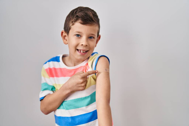 Молодой кавказский ребенок получает вакцину, показывающую руку с помощью пластыря, улыбаясь, показывая пальцем и рукой.  - Фото, изображение