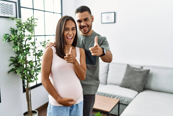 Młoda międzyrasowa para spodziewa się dziecka, dotykając brzucha w ciąży wskazując palcami do kamery ze szczęśliwą i zabawną twarzą. dobra energia i wibracje.  - Zdjęcie, obraz