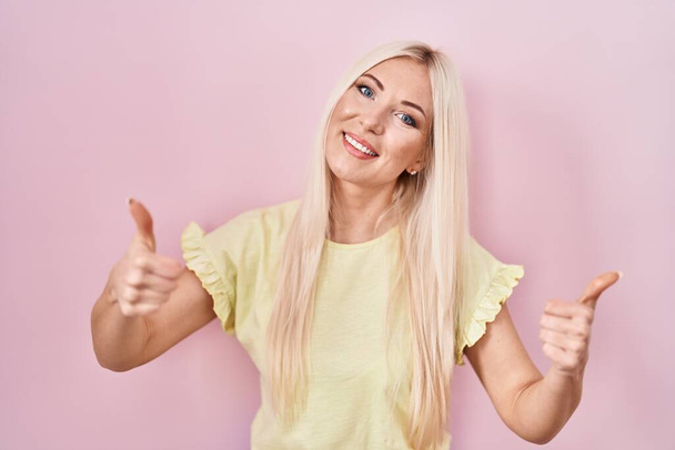 Caucasian woman standing over pink background approving doing positive gesture with hand, thumbs up smiling and happy for success. winner gesture.  - Foto, Imagem