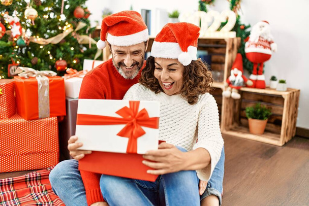 中世のヒスパニック夫婦はクリスマスの帽子をかぶっている。自宅で床の開口部の贈り物に座って. - 写真・画像