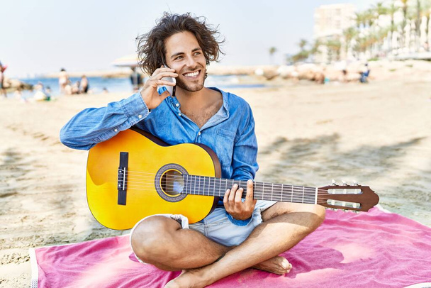 Young hispanic man playing guitar and talking on the smartphone sitting on sand at the beach. - Φωτογραφία, εικόνα