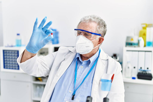 Hombre de pelo gris de mediana edad con uniforme científico hoding píldora en el laboratorio - Foto, imagen
