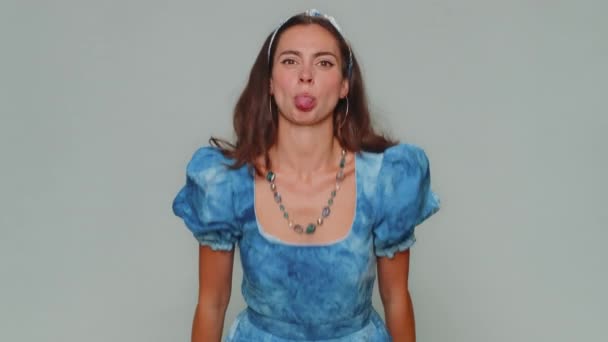 Cheerful funny bully young woman in dress. Adult girl showing tongue making faces at camera, fooling around, joking, aping with silly face, teasing isolated alone on gray studio background indoors - Imágenes, Vídeo