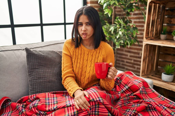 Mujer hispana joven sentada en el sofá tomando un café en casa haciendo cara de pez con labios, gesto loco y cómico. expresión divertida.  - Foto, Imagen