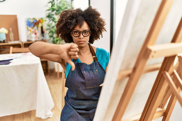 Beautiful african american woman with afro hair painting canvas at art studio with angry face, negative sign showing dislike with thumbs down, rejection concept  - Photo, Image