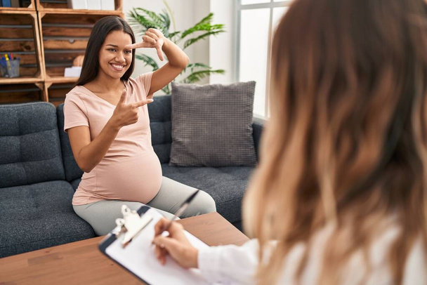 Jonge zwangere vrouw in therapie sessie glimlachend het maken van frame met handen en vingers met gelukkig gezicht. creativiteit en fotografie.  - Foto, afbeelding