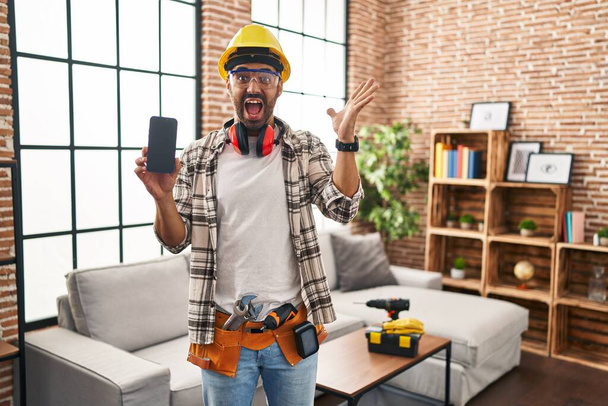 Junger hispanischer Mann mit Bart, der bei Renovierungsarbeiten zu Hause sein Smartphone hält und den Sieg mit glücklichem Lächeln und Siegerausdruck mit erhobenen Händen feiert  - Foto, Bild