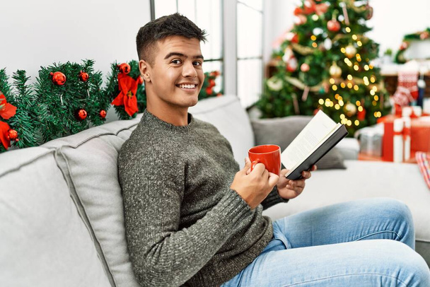Νεαρός Ισπανός διαβάζει βιβλίο πίνοντας καφέ καθισμένος στον καναπέ δίπλα στο χριστουγεννιάτικο δέντρο στο σπίτι - Φωτογραφία, εικόνα
