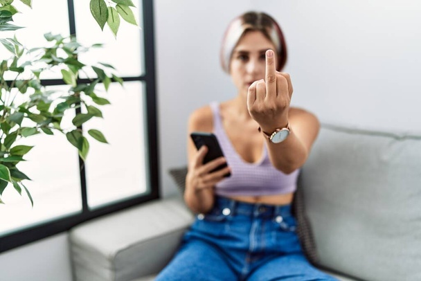 Young beautiful woman using smartphone typing message sitting on the sofa showing middle finger, impolite and rude fuck off expression  - Foto, Imagem