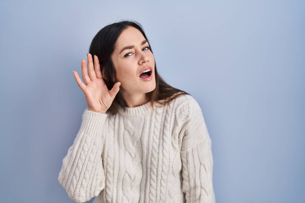 Young brunette woman standing over blue background smiling with hand over ear listening an hearing to rumor or gossip. deafness concept.  - Foto, afbeelding