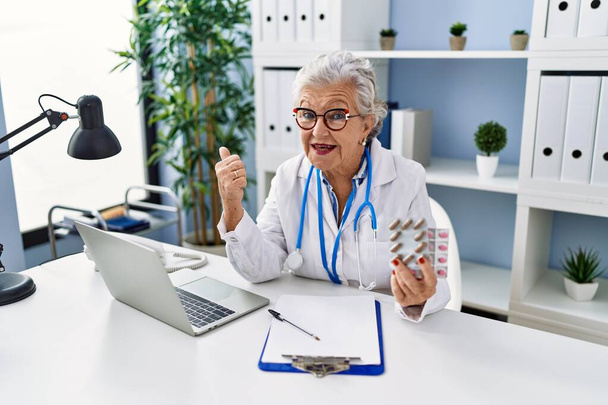 Mulher sênior com cabelos grisalhos vestindo uniforme médico segurando pílulas de prescrição apontando o polegar para o lado sorrindo feliz com a boca aberta  - Foto, Imagem