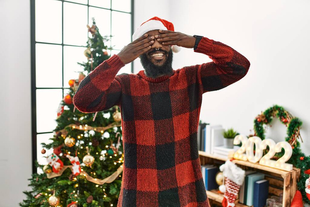 Αφροαμερικάνος που φοράει καπέλο Σάντα Κλάους και στέκεται δίπλα στο χριστουγεννιάτικο δέντρο καλύπτοντας τα μάτια με τα χέρια χαμογελαστά χαρούμενα και αστεία. τυφλή έννοια.  - Φωτογραφία, εικόνα