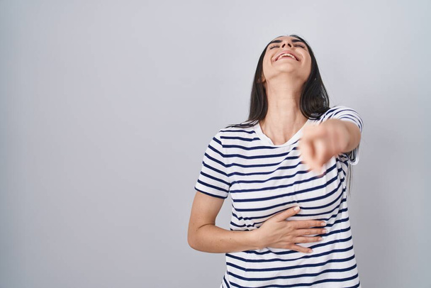 Young brunette woman wearing striped t shirt laughing at you, pointing finger to the camera with hand over body, shame expression  - Foto, Bild