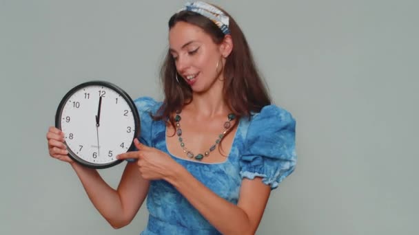 It is your time. Pretty young woman in blue princess dress showing time on wall office clock, ok, thumb up, approve, pointing finger at camera. Stylish female girl on gray studio background indoor - Imágenes, Vídeo