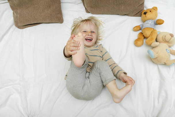 Niedliche kleine blonde kaukasische Kind Kleinkind Junge spielt, Spaß hat, auf dem Bett liegend, hebt die Beine zu Hause am sonnigen Morgen wacht in seinem Zimmer. Glückliche Kindheitsmomente. - Foto, Bild