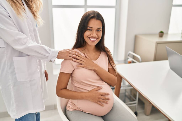 Young latin woman pregnant and doctor shake hands at clinic - Фото, изображение