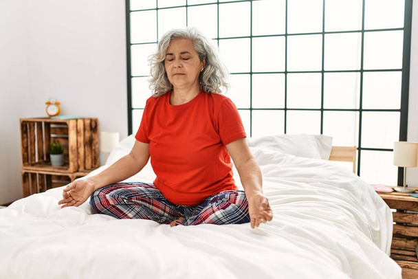 Жінка середнього віку сіроока займається вправами йоги, сидячи на ліжку в спальні
. - Фото, зображення