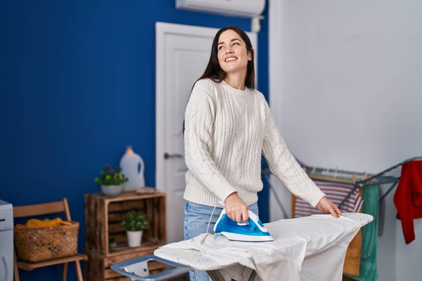 Young woman smiling confident ironing clothes at laundry room - 写真・画像