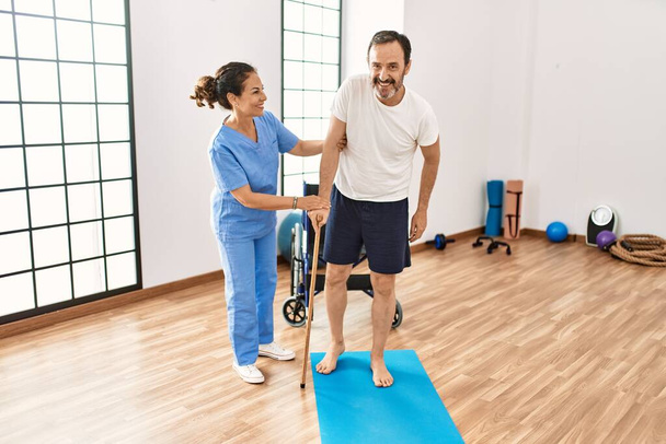 Uomo e donna di mezza età sorridenti fiduciosi di avere sessione di riabilitazione con bastone da passeggio alla clinica di fisioterapia - Foto, immagini