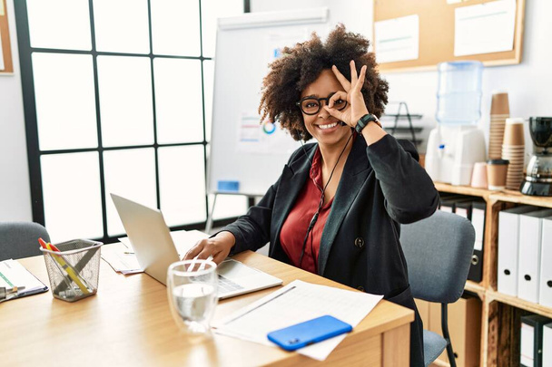 Mujer afroamericana con cabello afro trabajando en la oficina con auriculares de operador haciendo un buen gesto con la mano sonriente, ojos mirando a través de los dedos con la cara feliz.  - Foto, Imagen