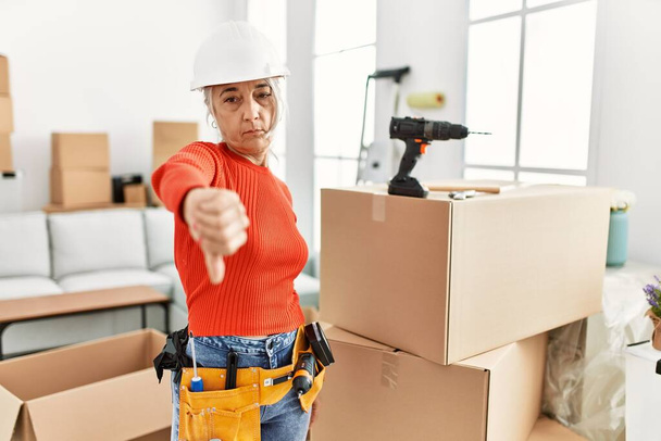 Middle age grey-haired woman wearing hardhat standing at new home looking unhappy and angry showing rejection and negative with thumbs down gesture. bad expression.  - Foto, afbeelding