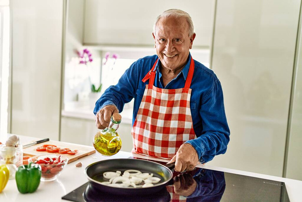 Senior man glimlachend zelfverzekerde gieten olie op koekenpan in de keuken - Foto, afbeelding