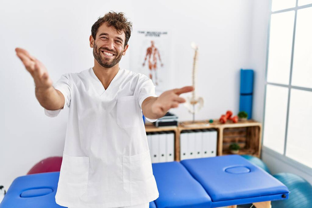 Jovem e bonito fisioterapeuta homem trabalhando na clínica de recuperação da dor sorrindo alegre oferecendo mãos dando assistência e aceitação.  - Foto, Imagem