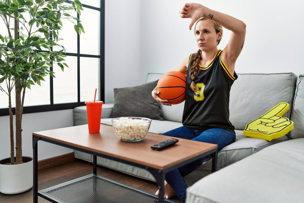 Young blonde woman holding basketball ball cheering tv game with angry face, negative sign showing dislike with thumbs down, rejection concept  - Foto, Imagen