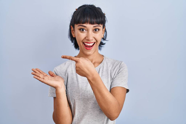 Fiatal spanyol nő visel alkalmi póló felett kék háttér meghökkent és mosolyog a kamera, miközben bemutatja a kezét, és ujjal mutogatott.  - Fotó, kép