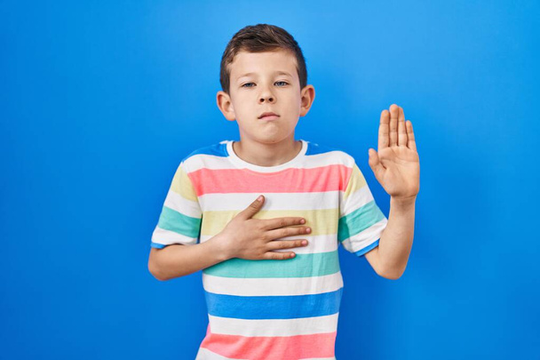 Young caucasian kid standing over blue background swearing with hand on chest and open palm, making a loyalty promise oath  - Fotografie, Obrázek