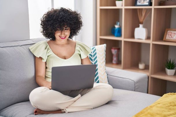 Young brunette woman with curly hair using laptop sitting on the sofa at home with a happy and cool smile on face. lucky person.  - Foto, Bild